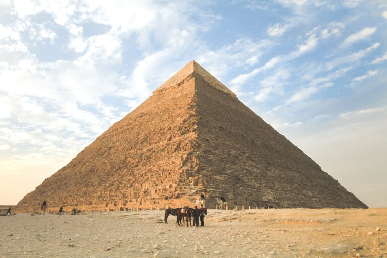a regal pyramid in Egypt sits behind a some people with horses and camels in the forefront.